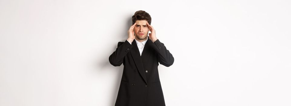 Image of businessman in black suit, touching head and looking dizzy, feeling painful headache, standing over white background.