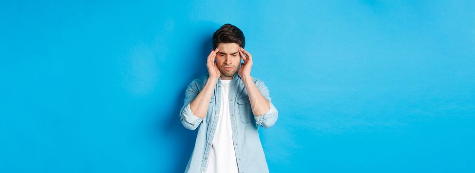 Man with hangover touching head and grimacing, having headache, standing against blue background.