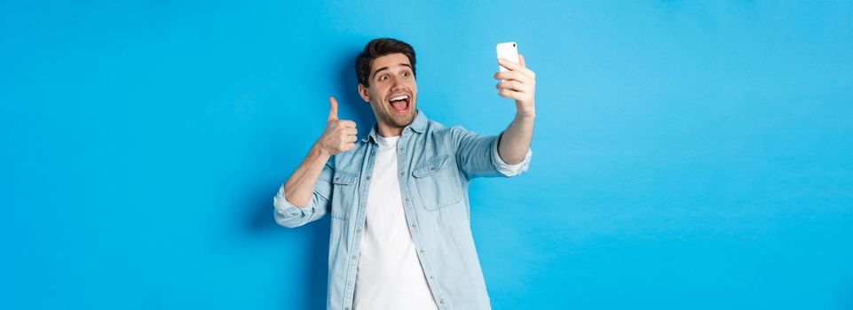 Happy man taking selfie and showing thumb up in approval on blue background, holding mobile phone.