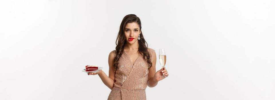 Party and celebration concept. Sexy woman in elegant dress, holding champagne and piece of cake, biting lip and looking seductive at camera.