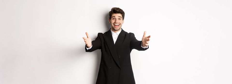 Portrait of happy attractive man in suit, hosting a party, reaching hands forward to greet you, want to hold something or hug, standing over white background.