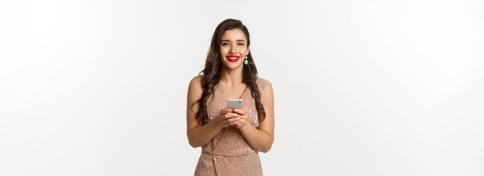 Christmas party and celebration concept. Happy young woman in elegant dress using mobile phone and smiling, shopping online, standing over white background.