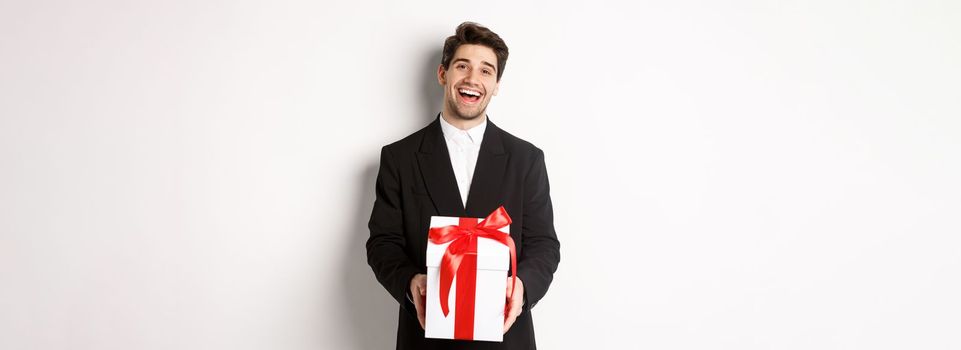 Concept of christmas holidays, celebration and lifestyle. Joyful handsome man in black suit, holding xmas gift and smiling, standing against white background.