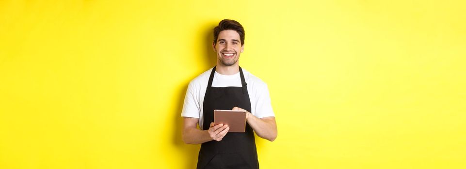 Handsome waiter taking orders, holding digital tablet and smiling, wearing black apron uniform, standing over yellow background.