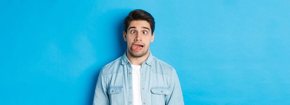 Close-up of young man making funny expressions, showing tongue and looking at camera, standing over blue background. Copy space