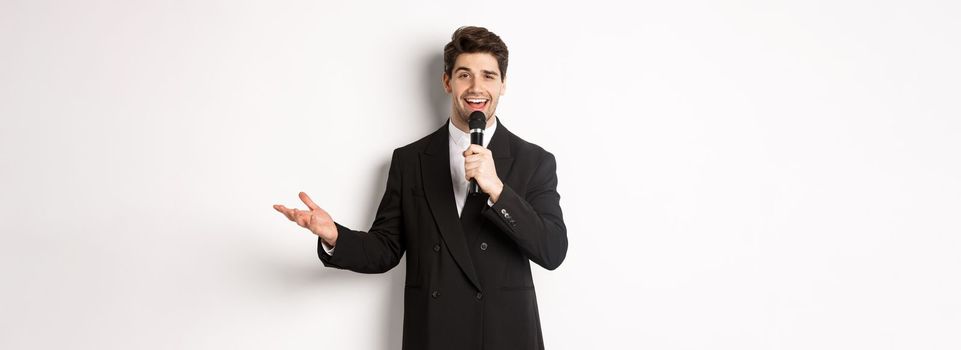 Portrait of handsome man in black suit singing a song, holding microphone and giving speech, standing against white background.