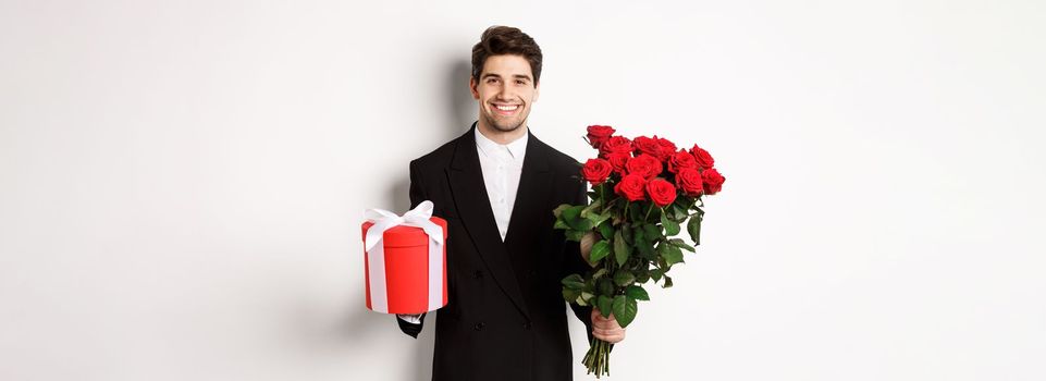 Concept of holidays, relationship and celebration. Handsome boyfriend in black suit, holding bouquet of red roses and a gift, wishing merry christmas, standing over white background.