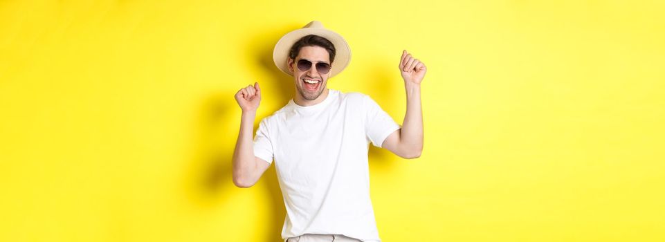 Tourism, travelling and holidays concept. Happy caucasian guy dancing and having fun on vacation, wearing sunglasses with straw hat, standing against yellow background.