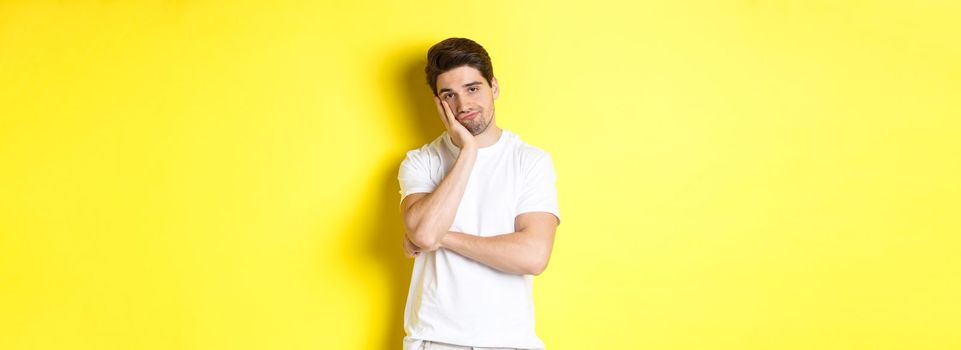 Bored and unamused man looking with indifference, standing in white clothes over yellow background.