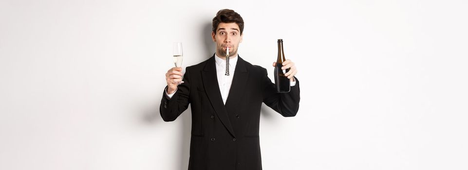 Concept of holidays, party and celebration. Portrait of handsome guy in black suit, raising bottle of champagne and glass, blowing a party whistle, having a birthday, standing over white background.