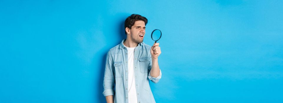 Young thoughtful guy looking through magnifying glass, reading something tiny, standing over blue background.