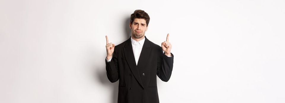 Portrait of reluctant and sad handsome man, complaining and pointing fingers up at something bad, looking jealous, standing over white background.