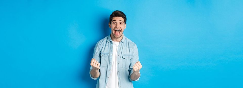 Excited handsome man winning, feeling lucky and rejoicing, celebrating victory and shouting yes, standing against blue background.