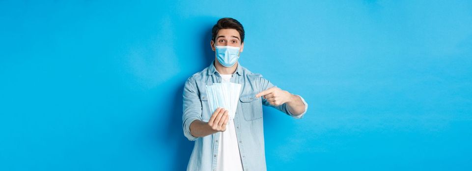Concept of coronavirus, quarantine and social distancing. Young man pointing at medical masks, preventing measures from covid-19, standing over blue background.