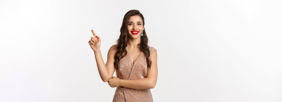 Beautiful woman in elegant dress on christmas party, pointing finger right at logo, smiling with red lips, standing over white background.