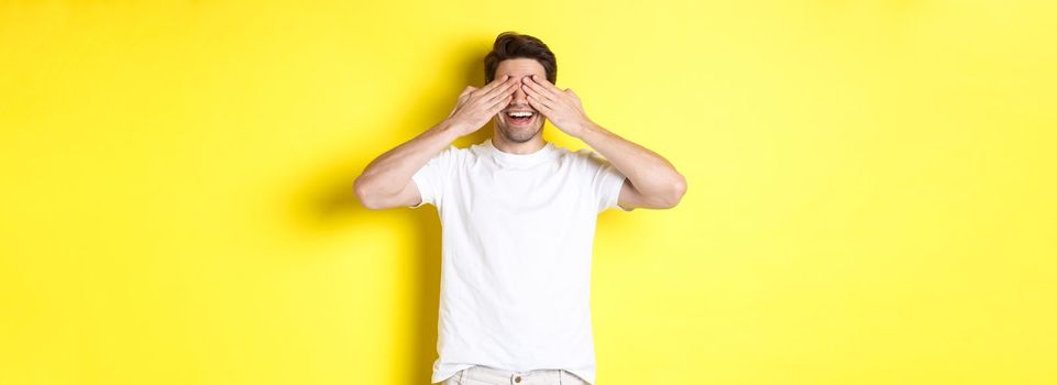 Happy man close eyes and waiting for surprise, smiling amused, standing over yellow background.