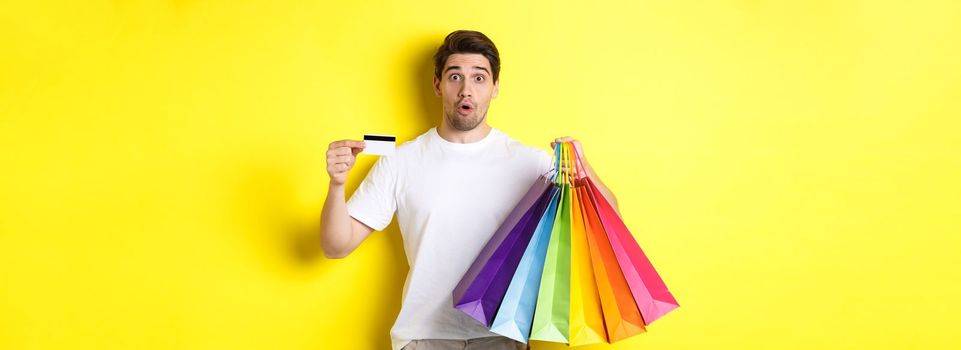 Excited man shopping on black friday, holding paper bags and credit card, standing against yellow background.