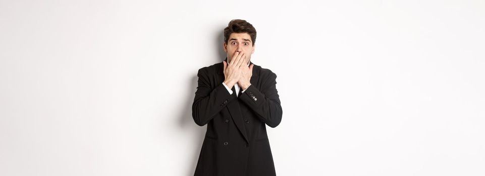 Scared man in formal suit, gasping and looking frightened at camera, standing against white background.
