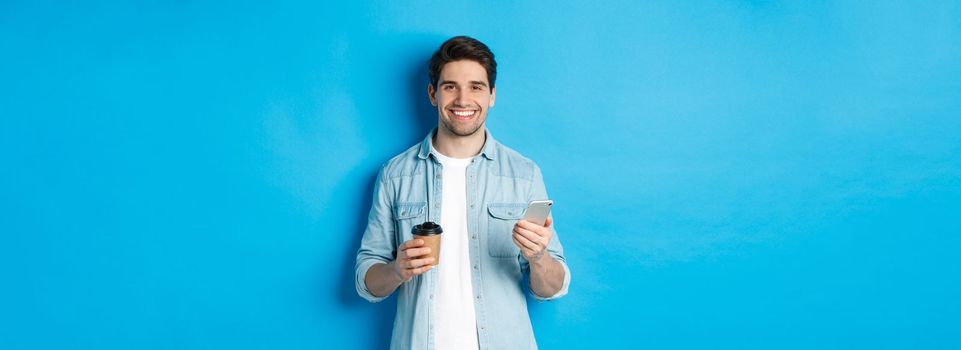 Young modern man drinking coffee and using mobile phone, smiling at camera, standing over blue background.