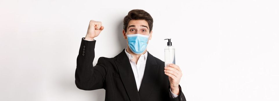 Concept of covid-19, business and social distancing. Close-up of happy man in trendy suit and medical mask, cheering and raising hand up, showing hand sanitizer, white background.