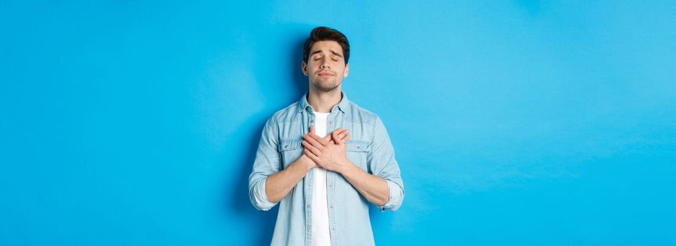 Portrait of dreamy and nostalgic guy in casual outfit, holding hands on heart and close eyes, daydreaming against blue background.