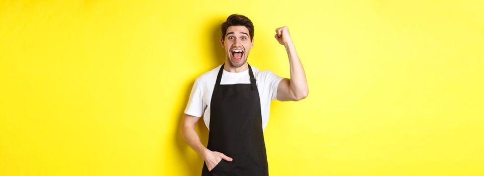 Cheerful seller making fist pump, rejoicing and triumphing, standing in black apron against yellow background.