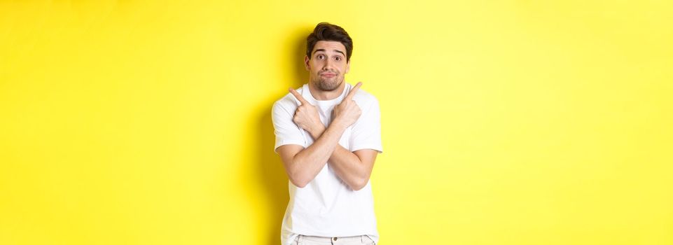 Indecisive man pointing fingers sideways, struggling to make decision, asking advice, standing over yellow background.