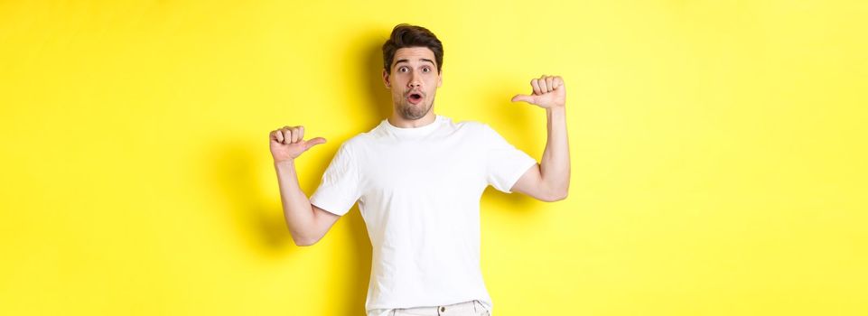 Surprised handsome guy pointing at himself, looking amazed, standing over yellow background. Copy space