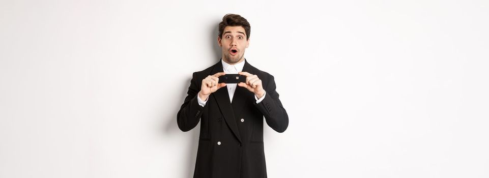 Portrait of handsome businessman in black suit, showing credit card and looking amazed, standing over white background.