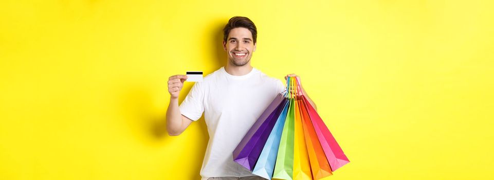 Young man shopping for holidays, holding paper bags and recommending bank credit card, standing over yellow background.