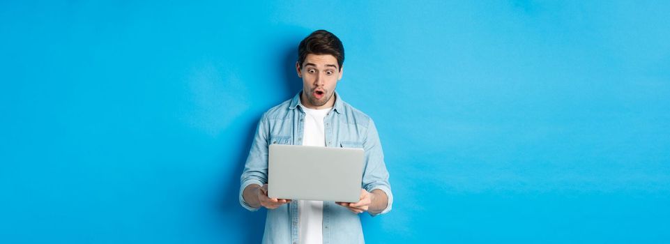 Impressed caucasian guy looking at laptop screen with amazement, checking out promo in internet, standing against blue background.