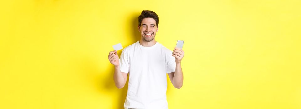 Happy young man shopping online in smartphone, holding credit card and smiling, standing over yellow background.