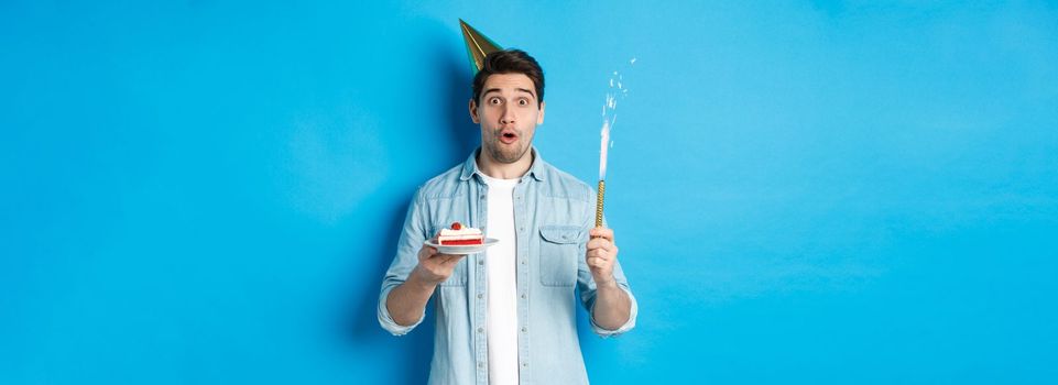 Happy young man celebrating birthday in party hat, holding b-day cake and smiling, standing over blue background.