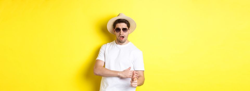 Handsome guy making finger gun shot and looking sassy, dressed for summer vacation in straw hat and sunglasses, standing against yellow background.