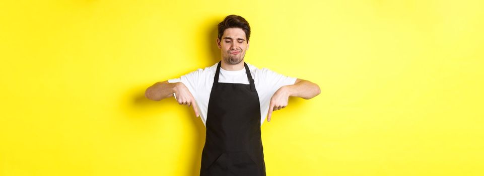 Doubtful barista complaining, pointing fingers down and grimacing displeased, standing in black apron against yellow background.