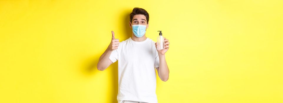 Concept of covid-19, quarantine and lifestyle. Satisfied young man in medical mask showing good hand sanitizer, thumbs up and recommending antiseptic, yellow background.