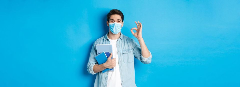 Education, covid-19 and social distancing. Guy student in medical mask looking happy, holding notebooks, showing ok sign, standing over blue background.