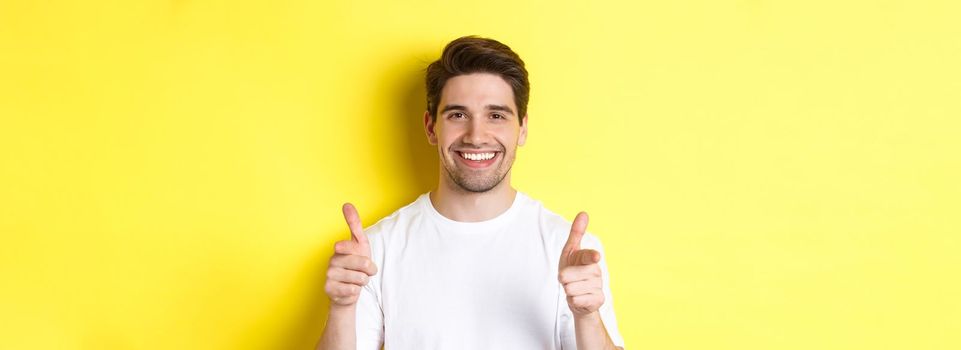Confident man pointing fingers at camera and smiling, praising you, standing over yellow background.