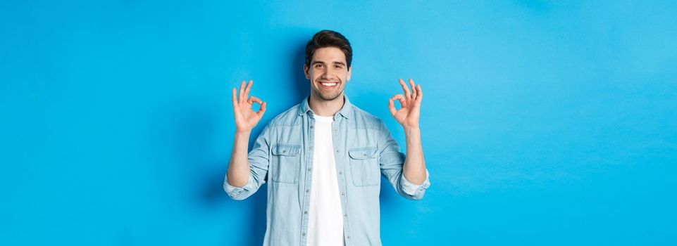 Confident smiling adult man, showing ok signs and looking pleased, like something, standing against blue background.
