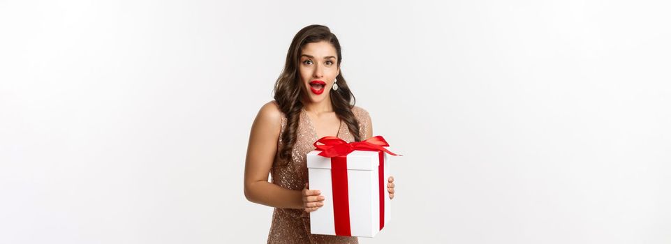 Merry Christmas. Image of beautiful woman in glamour dress receiving gift and looking surprised, celebrating New Year, standing over white background.