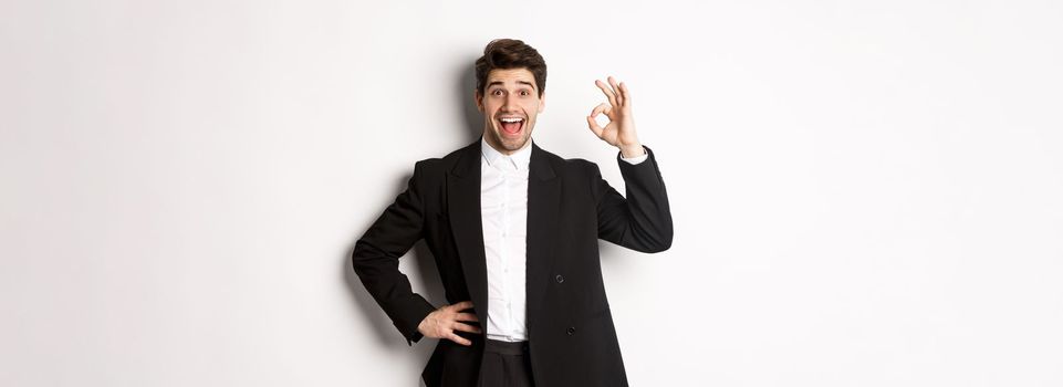 Concept of new year party, celebration and lifestyle. Portrait of pleased handsome guy in black suit, praise something good, showing okay sign in approval, standing over white background.