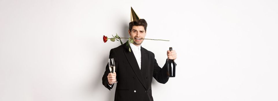 Romantic guy in trendy suit, celebrating and having a party, holding rose in teeth and champagne, standing over white background.