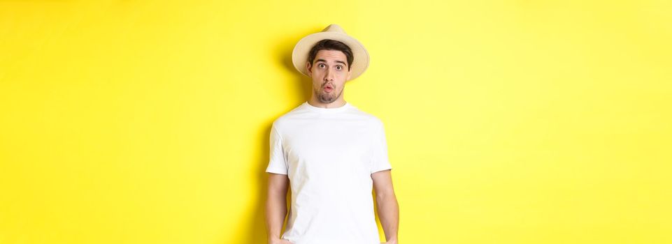 Confused guy traveller in straw hat looking puzzled, stare at camera, standing over yellow background. Copy space