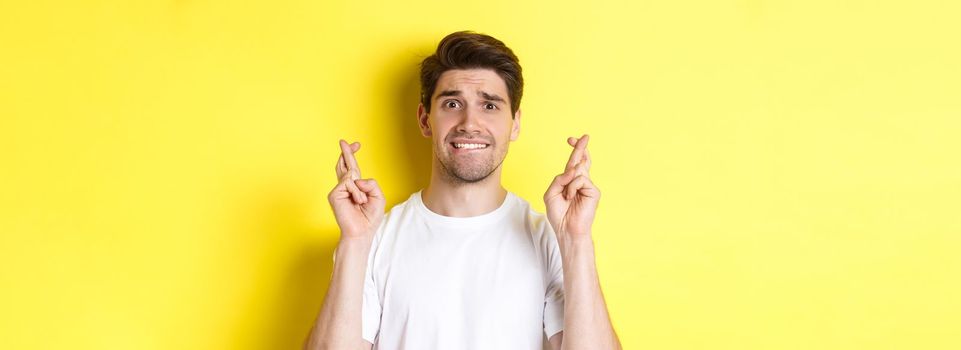 Close-up of nervous man making wish, holding fingers crossed and biting lip worried, standing over yellow background.