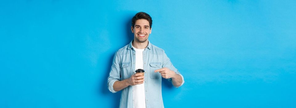 Smiling happy man pointing at paper cup with coffee, recommending cafe, standing over blue background.