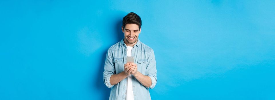 Image of handsome young man using mobile phone, texting on phone and looking pleased, standing over blue background.