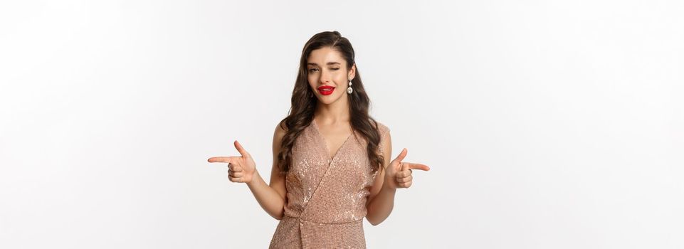 Concept of celebration, holidays and party. Beautiful and flirty woman with red lips, wearing glamour dress and winking, pointing fingers at camera, standing over white background.