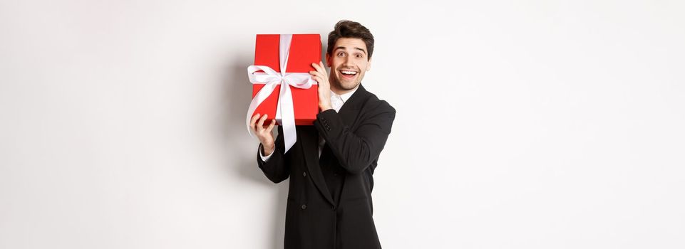 Concept of christmas holidays, celebration and lifestyle. Attractive man in black suit, holding new year gift and smiling, standing with a present over white background.