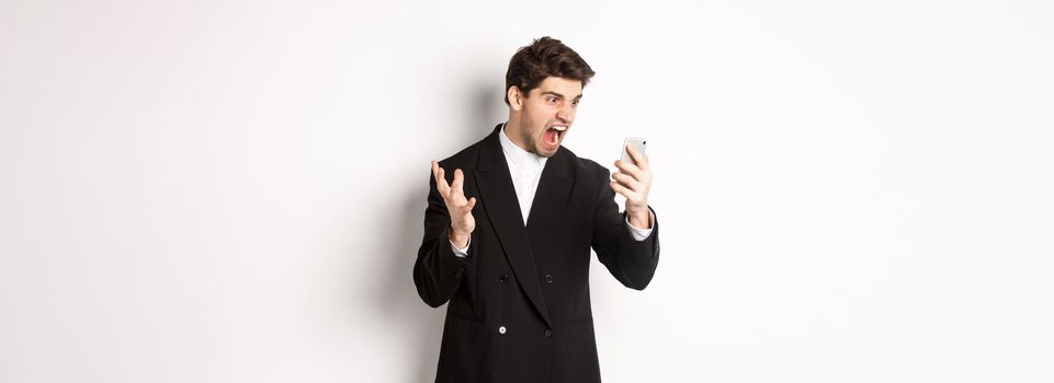 Portrait of angry businessman in black suit yelling at mobile phone, having an argument on video call, standing mad over white background.
