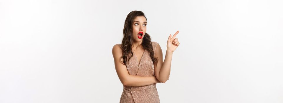 Christmas, holidays and celebration concept. Amazed fashionable woman in party dress and red lipstick, pointing and looking left at promo offer, standing against white background.